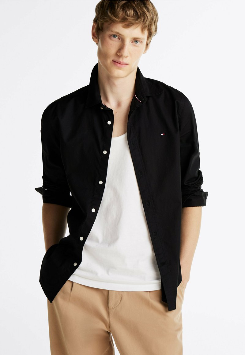 Chemise noire à manches longues avec une coupe décontractée, poignets à bouton, et un logo discret sur la poitrine. Portée par-dessus un débardeur blanc et un pantalon beige.