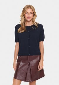 Marineblauwe korte mouw cardigan met een ronde halslijn en knoopsluiting, gecombineerd met een bordeauxleren geplooide rok. Soepele textuur.