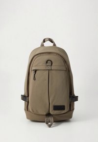 Vaaleanruskea kangasreppu, jossa pyöreä yläosa, etutaskun vetoketju, sivulla solki ja näkyvä "HUGO"-logo.