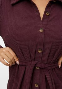 Geribbelde bordeaux kleurige jurk met knoopsluiting aan de voorkant, kraag en een gestrikt middel. Bevat bruine knopen en een gestructureerde stof.