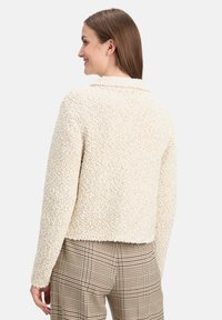 Veste texturée couleur crème avec un col et des manches longues, portée sur un pantalon à carreaux. Le tissu semble doux et duveteux.