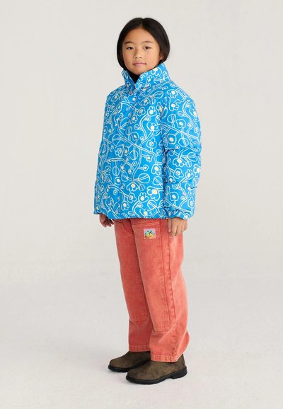 Enfant portant une doudoune bleue à motif floral, un pantalon corail avec un patch et des bottes marron foncé, debout devant un fond uni.
