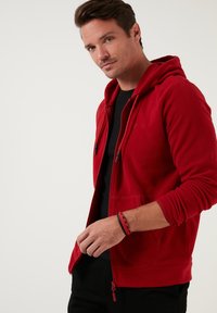 Crvena zip-up dukserica napravljena od mekanog materijala. Ima kapuljaču sa vezicom, prednje džepove i elastikane manžetine. Nosi se preko crne majice.