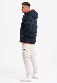 Chaqueta de plumas azul marino con capucha, forro naranja en contraste, textura acolchada, corte estándar; combinada con pantalones cargo claros y zapatillas blancas.