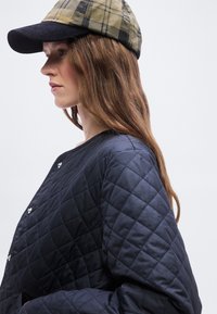 Γυναίκα με μακριά καστανά μαλλιά, που φοράει καπέλο κάρο σε καφέ και μαύρο χρώμα και μπλε σκούρο καπιτονέ μπουφάν με κουμπιά πάτημα, κοιτάζει προς τα αριστερά.