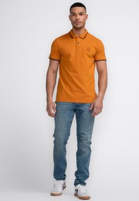 Oranžové polo tričko s černým lemem, dvěma knoflíky a logem na hrudi. Nosené s světle modrými džíny a bílými teniskami s černými akcenty.