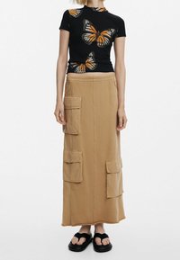 Camiseta negra de manga corta con estampado de mariposas en naranja y blanco, combinada con una falda maxi color marrón claro que tiene bolsillos laterales y un ajuste relajado.