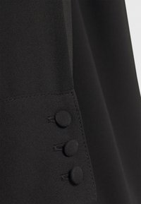Gros plan d'un poignet de vêtement noir avec trois boutons recouverts de tissu en rangée verticale et des coutures visibles le long du bord.