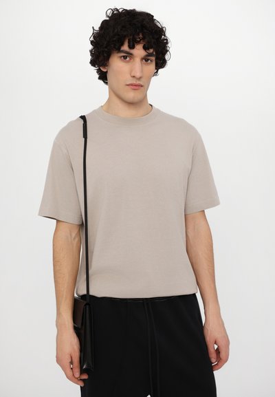 Beige cotton T-shirt with short sleeves and a round neckline, paired with black pants. Features a slim black crossbody strap and pocket.