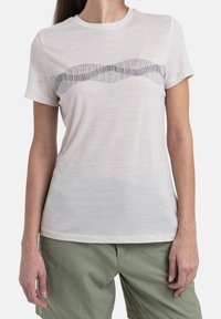 Ljusbeige t-shirt med kort ärm, mjuk textur, med ett vingligt mönster i mörkgrått över bröstet. Kombineras med gröna byxor.