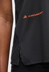 Gros plan sur un vêtement de sport noir perforé avec le logo adidas orange et la marque H.Koumori à côté du bras d'une personne.