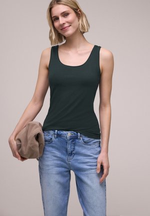 Kobieta w ciemnozielonym topie bez rękawów i jasnoniebieskich dżinsach, trzyma beżową złożoną kurtkę w lewej ręce.