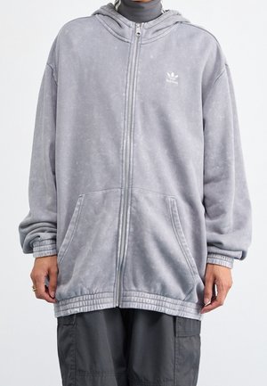 Person iført en lysegrå Adidas hættetrøje med lynlås og forlommer samt mørkegrå cargobukser, hænderne hvilende ved siderne.