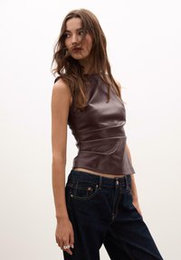 Brun læder ærmeløs top med en tætsiddende design og sidesøm; bæres med mørkeblå jeans med synlige syningsdetaljer.