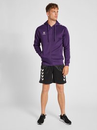 Felpa viola con zip e cappuccio, accompagnata da pantaloni corti neri con motivi a zig-zag. Indossata con scarpe sportive nere, design minimalista.