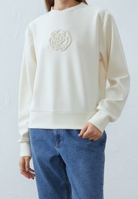 Sudadera de color crema fabricada con un tejido suave, que presenta un patrón de rosas blancas texturizadas en el pecho y puños y dobladillo acanalados.