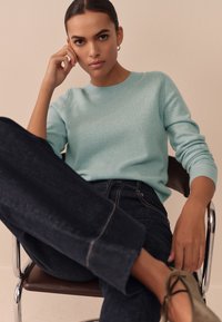 Lichtblauwe gebreide trui met een ronde hals, gecombineerd met donkerblauwe spijkerbroek. Het model draagt olijfgroene schoenen en zit op een chromen stoel.