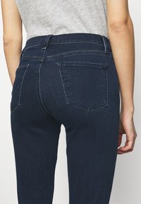 Jeans de mezclilla azul oscuro con una cintura de altura media, dos bolsillos traseros y detalles de costura sutiles, que cuentan con una textura suave y un ajuste entallado.