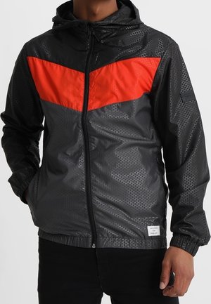 Veste coupe-vent noire avec une surface texturée, présentant une bande en chevron orange vif sur la poitrine et une capuche ajustée.