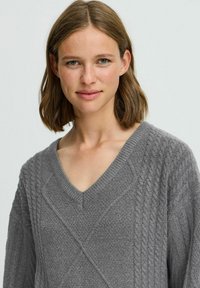 Grauer Strickpullover mit V-Ausschnitt, strukturiertem Muster und Kabeldetails sowie gerippten Rändern. Weiches Material erhöht den Tragekomfort.
