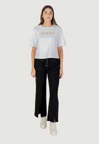 Witte cropped t-shirt met "ARMANI" in gouden letters, gecombineerd met zwarte wijde broek met uitsneden en een strik in de taille. Witte sneakers maken de look compleet.