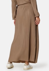 Lange, geplooide jurk in een taupe kleur met een bijpassende riem. Heeft lange mouwen en is gecombineerd met witte sneakers. Zachte textuur.