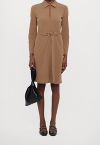 Robe marron à manches longues avec un décolleté en forme de goutte et une taille ceinturée. Accompagnée d'une pochette noire et de chaussures plates marron avec des détails à brides.