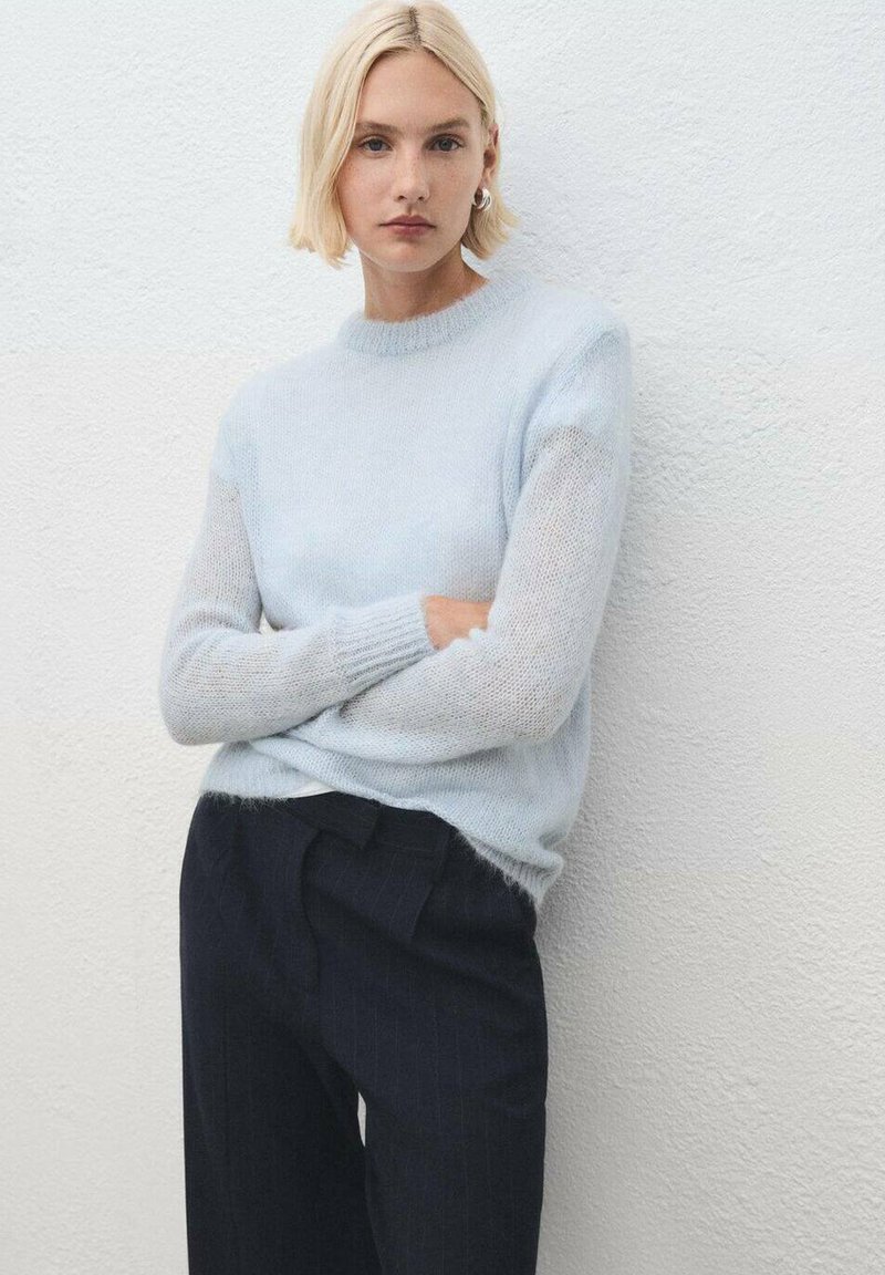 Maglione lavorato a maglia blu chiaro con maniche testurizzate, indossato sopra pantaloni scuri a righe. Il materiale sembra morbido e leggermente trasparente.