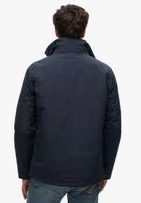 Marineblauwe waterdichte jas met hoge kraag, gladde textuur en verborgen naaddetails, gecombineerd met lichtblauwe denim jeans.