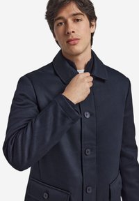 Marineblauer Mantel aus strukturiertem Stoff mit einem Kragen, Knopfleiste und zwei Vordertaschen. Maßgeschneiderte Passform.