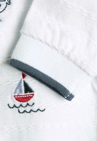 Tela de punto blanca con rayas texturizadas. Presenta un puño gris y un detalle de velero bordado en rojo, negro y plata por encima de líneas onduladas.