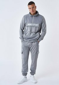 Lys grå træningsdragt med hætte. Har en frontlomme, elastiske manchetter og bukser med lynlåslommer. Følger med hvide sneakers.
