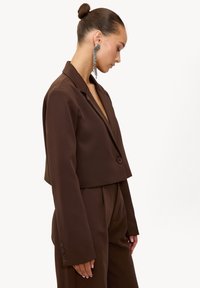 Femme aux cheveux attachés en chignon portant un blazer court marron foncé et un pantalon assorti, accessoirisée de longues boucles d'oreilles argentées pendantes.