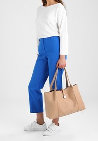 Kvinna i en vit långärmad topp, blå cropped byxor, vita sneakers, som håller en stor beige tygväska i handtagen.