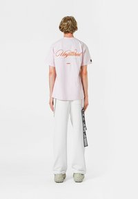 Camiseta de algodón rosa claro con "Desatado" impreso en naranja en la parte posterior, combinada con pantalones blancos de corte holgado y calzado gris.