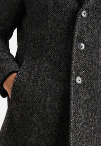 Donkergrijze wollen jas met een gestructureerd patroon, drie zichtbare zwarte knopen en een hand die gedeeltelijk in de linkerzijzak is gestoken.