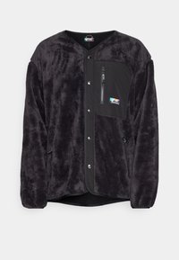 Giacca in pile nera e pelosa con bottoni a pressione, tasca sul petto con zip, tasche laterali e un piccolo logo colorato a forma di montagna. 