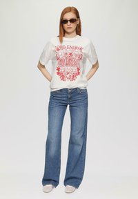 Witte katoenen t-shirt met rode bloemenprint en de tekst "GOEDE ENERGIE," gecombineerd met blauwe wijde jeans en instap schoenen. Accessoires zijn onder andere een donker zonnebril.