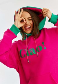Fioletowa bluza z kapturem z zielonymi akcentami, z napisem "Ciao!" w pogrubionym tekście. Posiada przednią kieszeń oraz sznurki z metalowymi końcówkami.