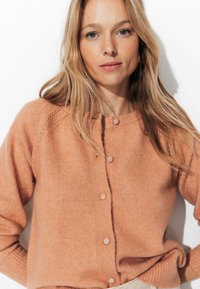 Cardigan en tricot couleur pêche avec un décolleté arrondi, des boutons de fermeture et des poignets côtelés. Présente une finition texturée et de subtils motifs en œillets.