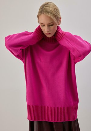 Femme ajustant le col d’un pull en grosse maille rose vif oversize sur un fond gris clair uni.