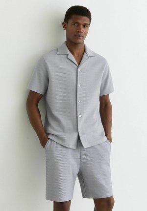 Muž stojaci pri bielej stene, oblečený v svetlosivej krátkorukávovej košeli na gombíky a v rovnakej textúrované krátke nohavice, s rukami vreckách.
