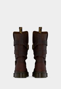 Ζευγάρι σκούρα καφέ δερμάτινα μποτάκια αστραγάλου με λουριά και αγκράφες και χοντρές σόλες από καουτσούκ με κίτρινα ραφές, από πίσω όψη.