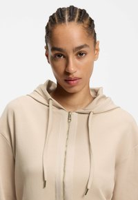 Sudadera con capucha beige con cremallera, capucha grande, cordones y cremallera dorada. Textura suave y ajuste relajado, con detalles de costuras verticales.