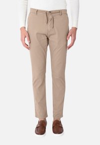 Pantaloni in cotone elasticizzato beige con vita regolabile con cordino, vestibilità slim e due tasche laterali, abbinati a scarpe marroni senza lacci.