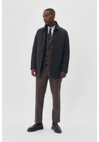 Cappotto impermeabile nero con bottoni lunghi sopra un completo marrone, una camicia bianca e una cravatta a righe; pantaloni grigi e scarpe nere completano il look.