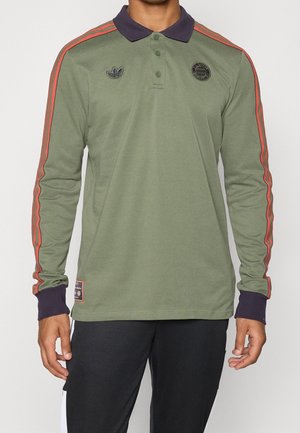 Mann trägt olivgrünes Langarm-Poloshirt mit roten Streifen an den Ärmeln, marineblauem Kragen und Bündchen sowie schwarze Hose vor weißem Hintergrund.
