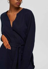 Robe chemise bleu marine foncé avec un col en V, des manches longues et une taille nouée. Conçue dans un tissu lisse avec une coupe décontractée et des poches latérales.