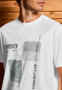 Bílý tričko se geometrickým grafickým designem v šedé a černé barvě s textem "ELEVATE YOUR STYLE" a "CONCEPT LAB" vytištěným na přední straně.
