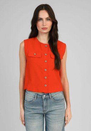 Femme aux longs cheveux foncés portant un haut sans manches orange vif boutonné et un jean bleu clair, debout devant un fond gris.